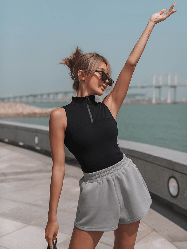 Woman wearing sporty black sleeveless top with gray shorts, stylish sunglasses, posing confidently outdoors.